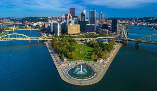 Photograph of Pittsburgh taken from the sky off the Point State Park fountain. From this angle, it is framed by the Fort Duquesne and Fort Pitt Bridges. The foundtain is at a point where the Ohio River splits under the two bridges. Behind the fountain is a grassy area (Fort Duquesne> and then the bridges connecting and finally the city skyline. It is a bright and sunny day.